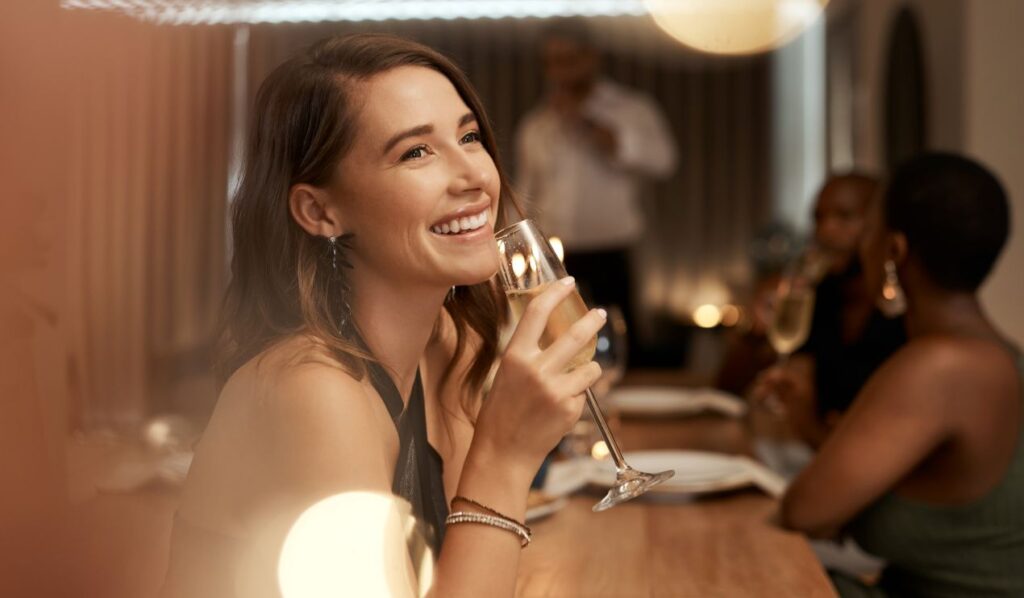 Lächelnde Frau hält ein Glas Sekt bei einer Feier, umgeben von stimmungsvoller Beleuchtung und geselligen Gästen im Hintergrund.